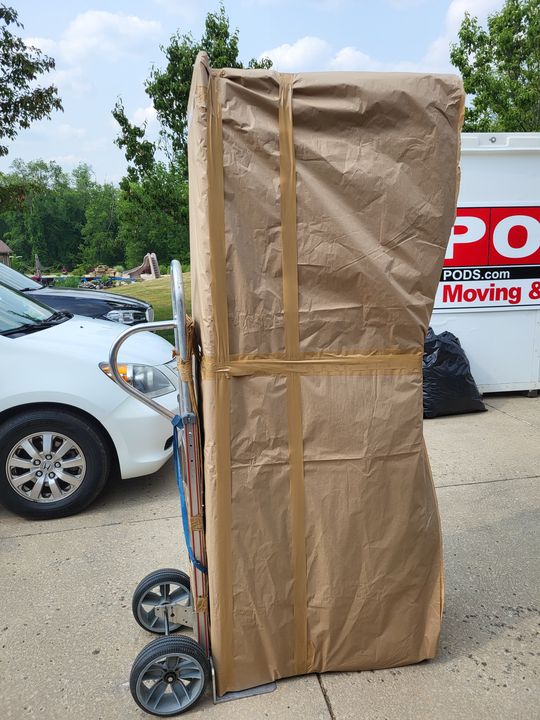 Tall paper-wrapped item on a hand truck beside a PODS container (Labor only moving) by Moving Pros CLE